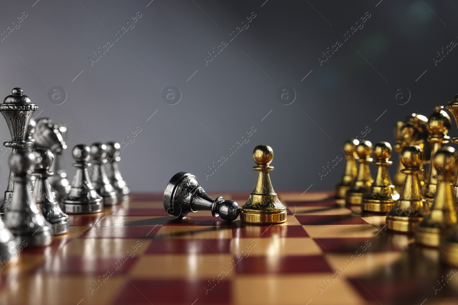 Photo of Golden pawn near fallen silver one and other chess pieces on chessboard against grey background, closeup. Competition concept