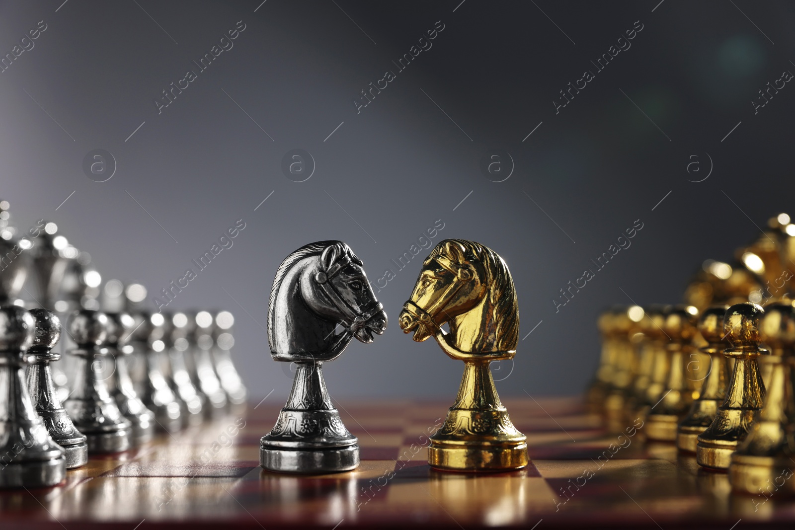 Photo of Golden and silver knights near other chess pieces on chessboard against grey background, closeup. Competition concept