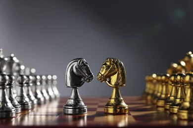 Photo of Golden and silver knights near other chess pieces on chessboard against grey background, closeup. Competition concept