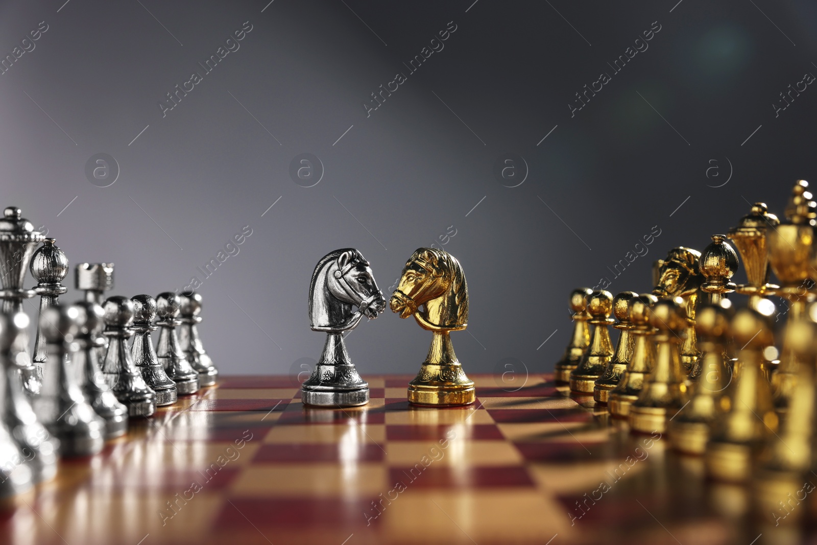 Photo of Golden and silver knights near other chess pieces on chessboard against grey background, closeup. Competition concept