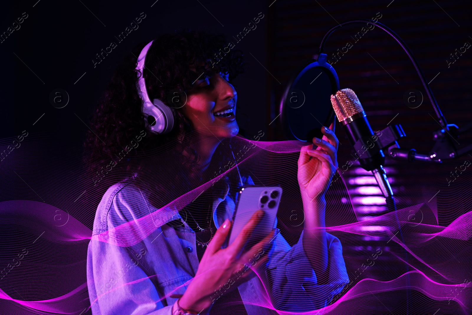 Image of Vocalist with smartphone and headphones singing into microphone in professional record studio. Sound waves