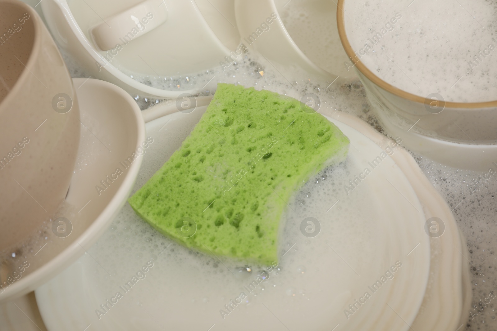 Photo of Green sponge, dishes and foam in sink, closeup