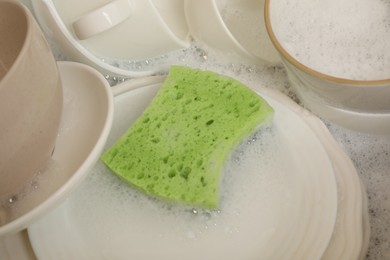 Photo of Green sponge, dishes and foam in sink, closeup