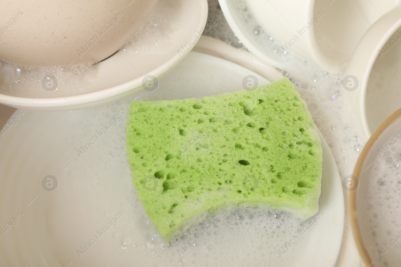 Photo of Green sponge, dishes and foam in sink, closeup