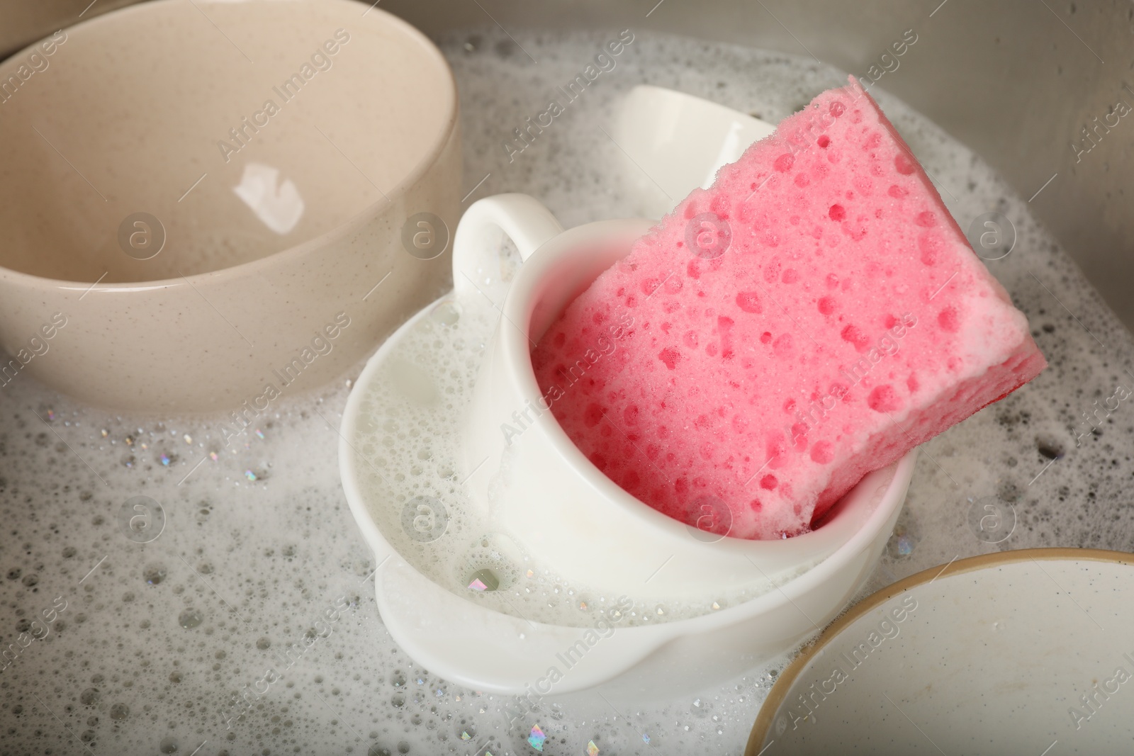 Photo of Pink sponge, dishes and foam in sink, closeup