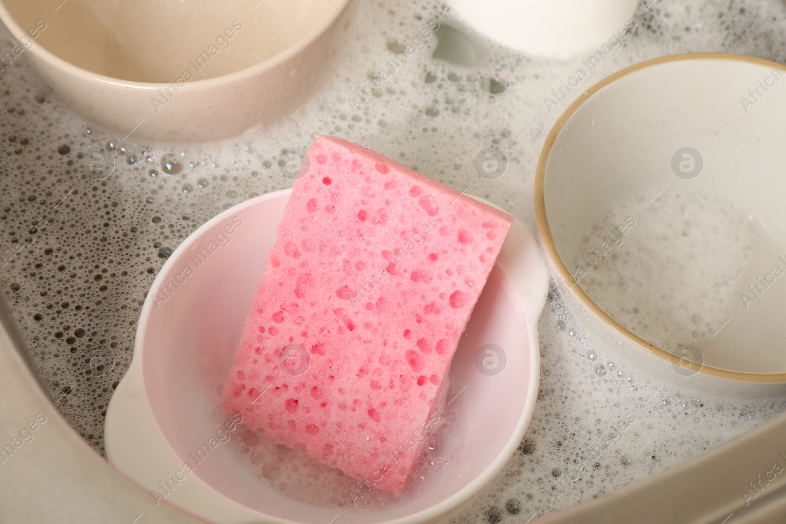 Photo of Pink sponge, dishes and foam in sink, closeup