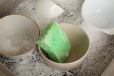 Photo of Green sponge, dishes and foam in sink, closeup