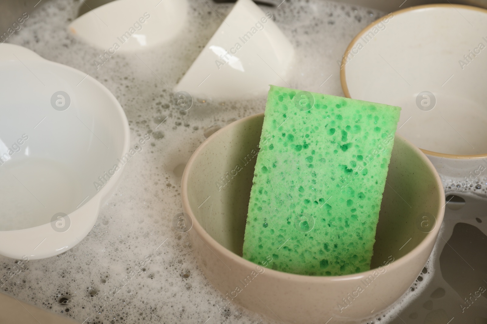 Photo of Green sponge, dishes and foam in sink, closeup