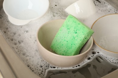 Photo of Green sponge, dishes and foam in sink, closeup