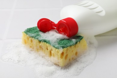 Photo of Sponge, foam and bottle of dish soap on white tiled table, closeup