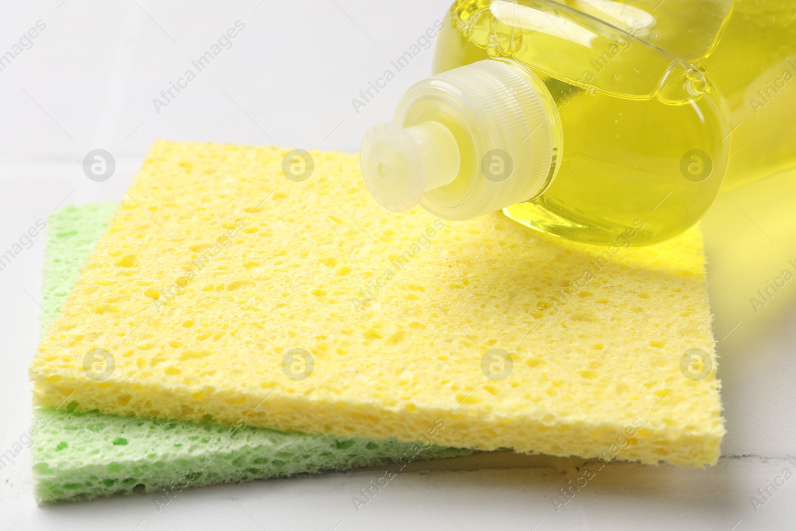 Photo of Sponges and bottle of dish soap on white tiled table, closeup