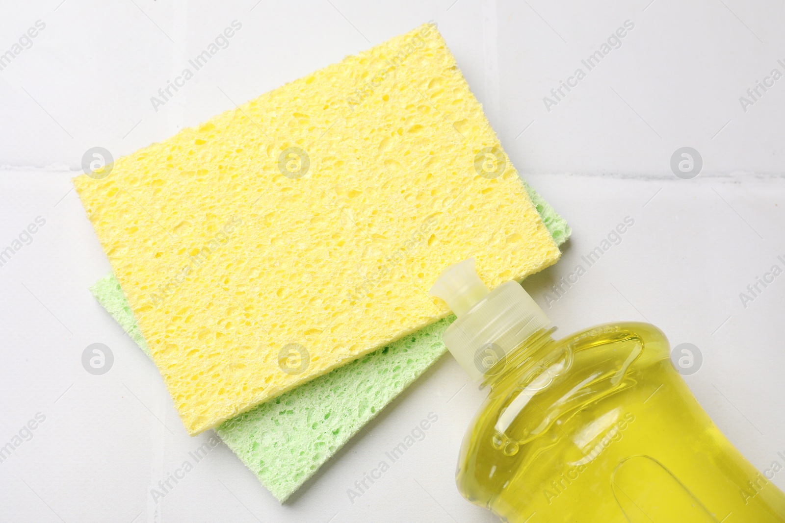 Photo of Sponges and bottle of dish soap on white tiled table, flat lay