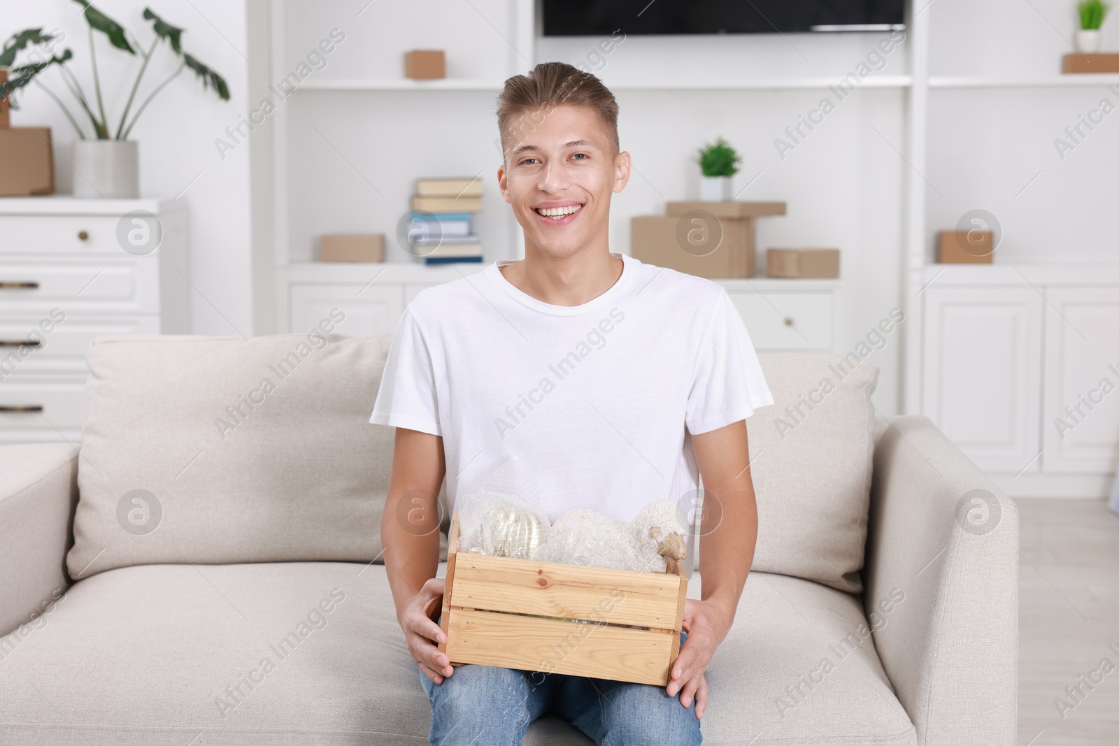 Photo of Happy man holding wooden crate with stuff on sofa in new apartment. Housewarming party