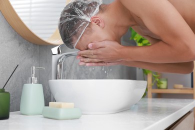 Photo of Man wearing shower cap washing his face in bathroom