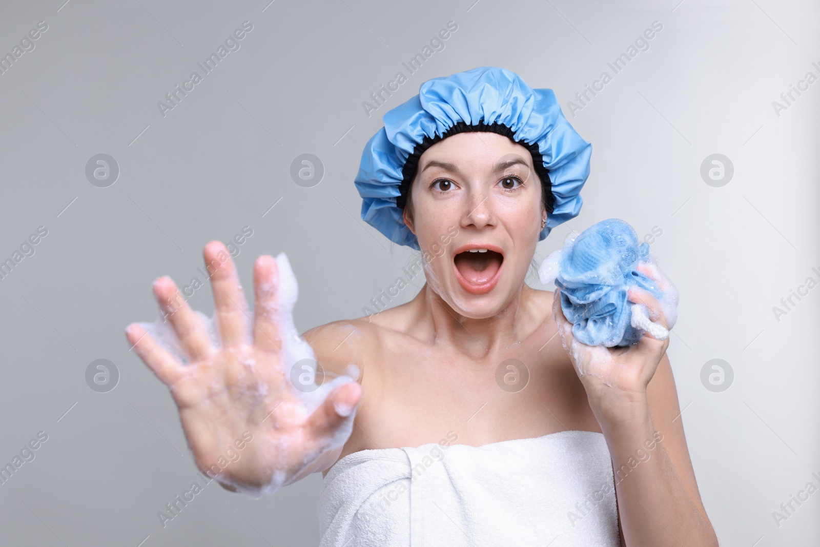 Photo of Emotional woman with shower cap and mesh sponge making stop gesture on grey background