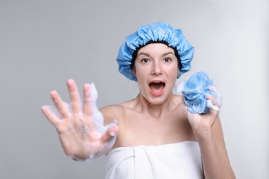 Photo of Emotional woman with shower cap and mesh sponge making stop gesture on grey background