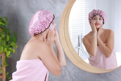 Photo of Woman with shower cap applying face cream near mirror in bathroom