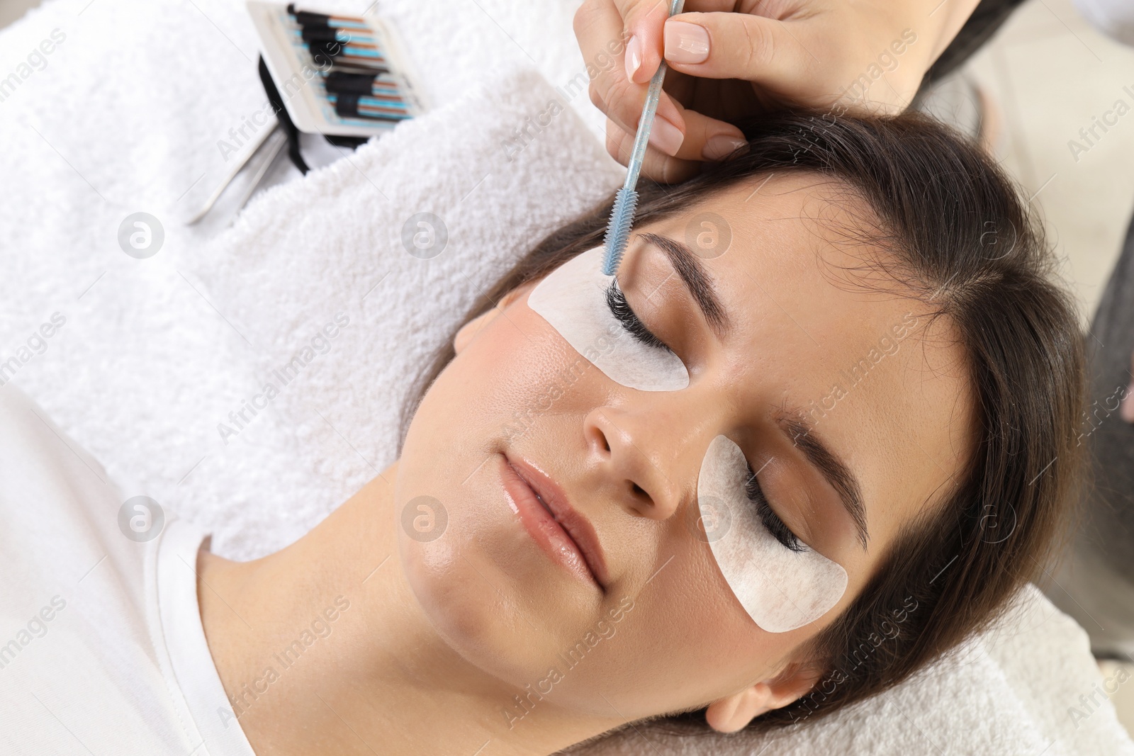 Photo of Esthetician brushing woman's lash extensions during procedure in beauty salon, closeup