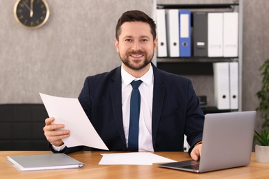 Photo of Banker holding document and working with laptop at wooden table in office