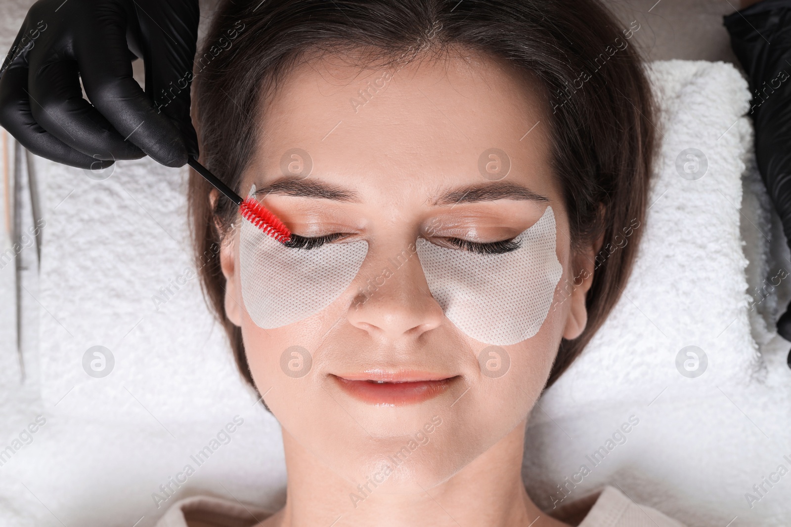 Photo of Eyelash extension procedure. Esthetician combing woman's lashes with brush in beauty salon, top view