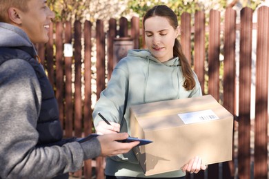 Photo of Woman signing papers while receiving parcel from postman outdoors