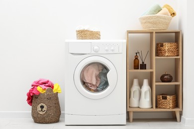 Photo of Wicker basket full of laundry, washing machine and detergents in bathroom