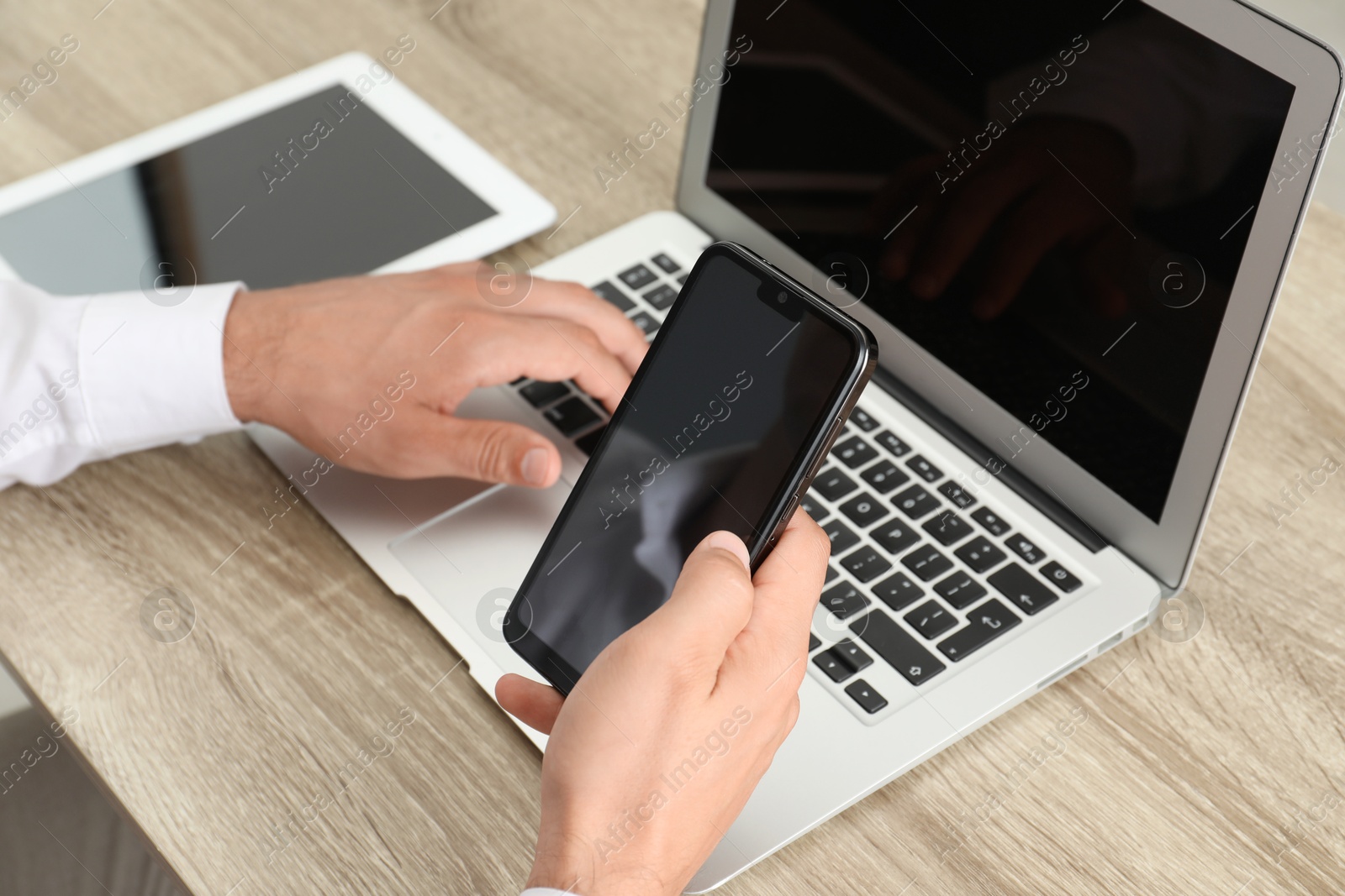 Photo of Businessman with smartphone using laptop at wooden table, closeup. Modern technology