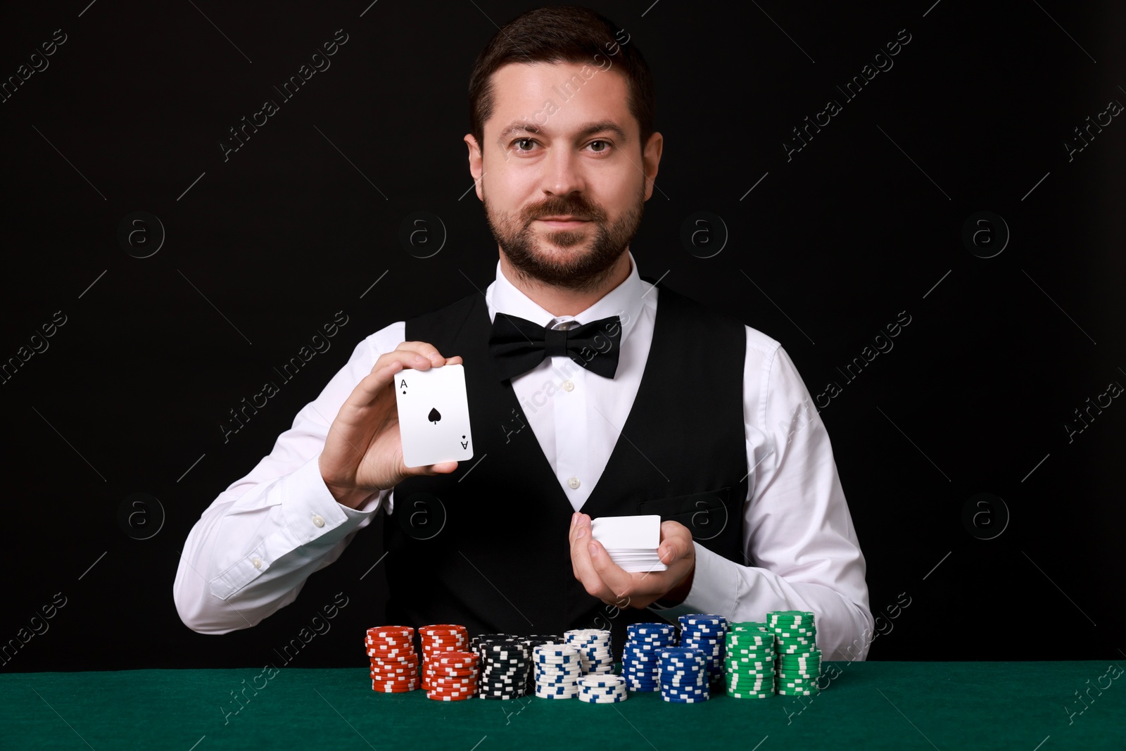 Photo of Professional croupier with playing cards at gambling table against black background