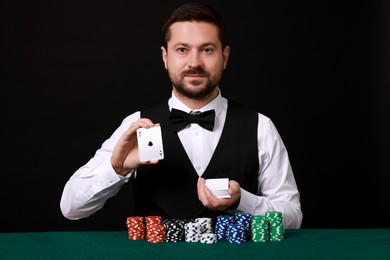 Photo of Professional croupier with playing cards at gambling table against black background