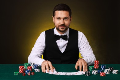 Photo of Professional croupier with playing cards at gambling table against dark yellow background