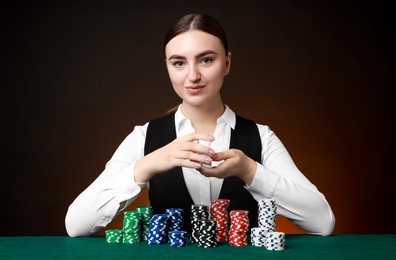 Professional croupier with casino chips and playing cards at gambling table on color background