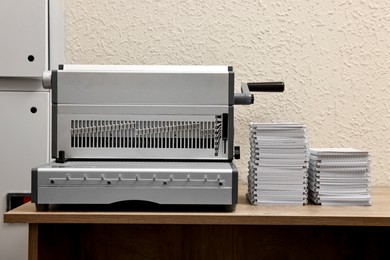 Photo of Modern binding machine and stacks of notebooks on wooden table indoors