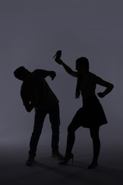 Domestic violence. Silhouette of woman with high-heeled shoe threatening her husband on grey background