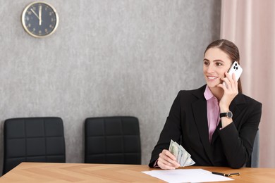 Photo of Banker with dollar banknotes talking on smartphone at wooden table in office