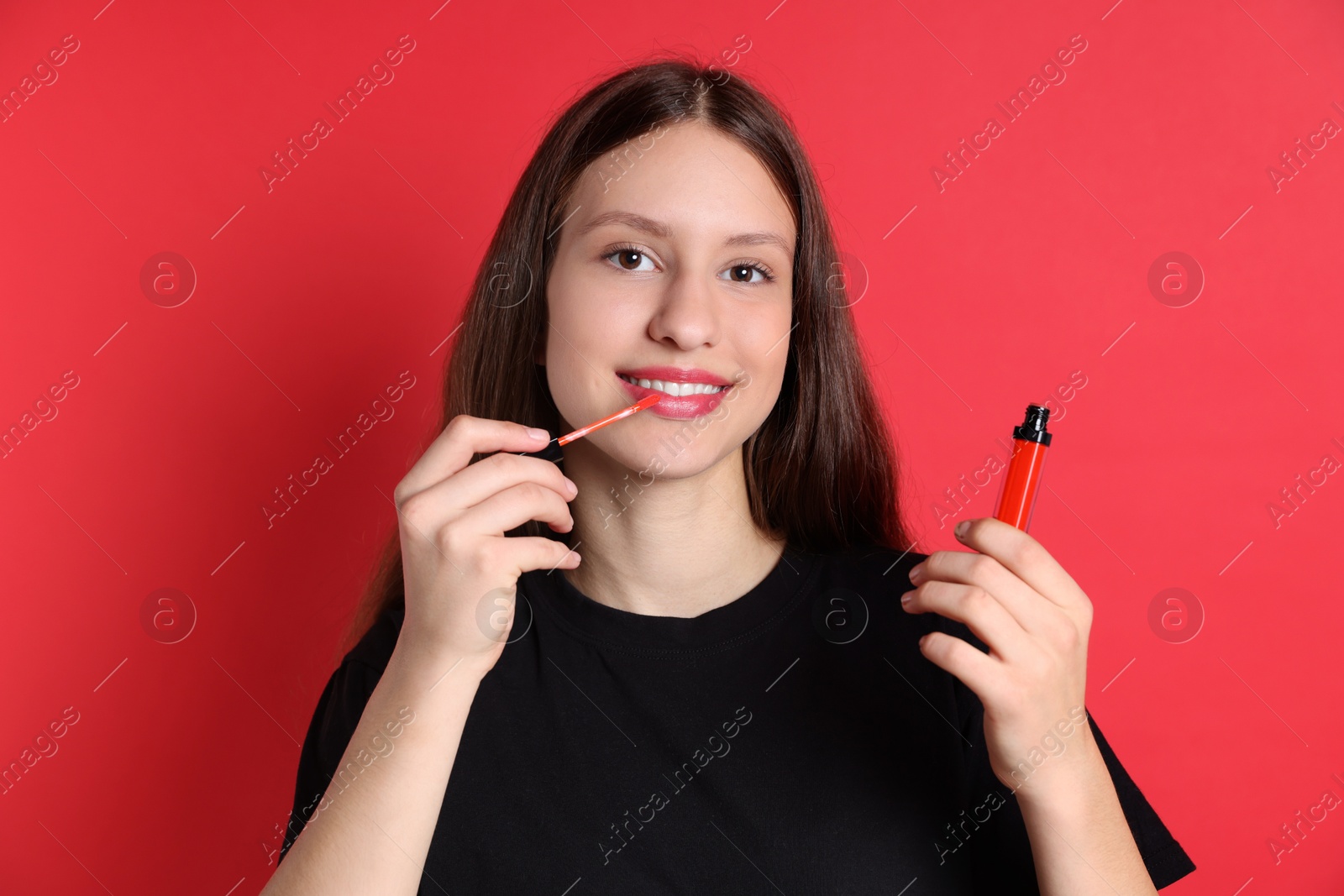 Photo of Smiling teenage girl applying lip gloss on red background