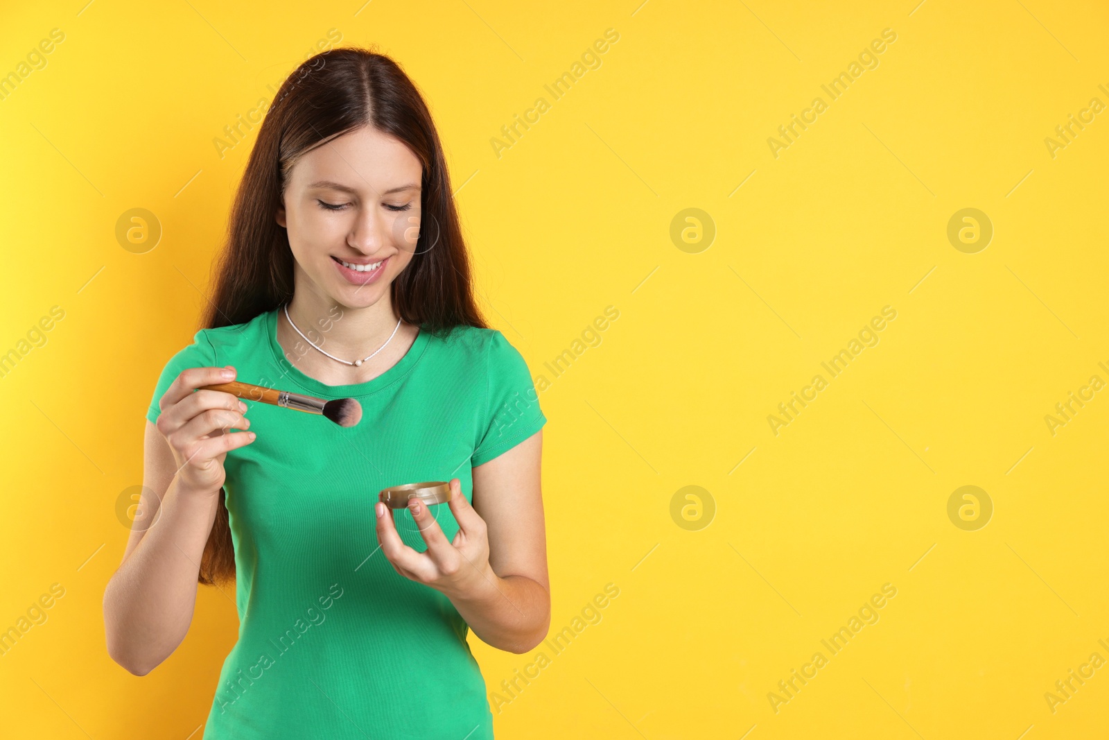 Photo of Smiling teenage girl with blusher and makeup brush on yellow background. Space for text