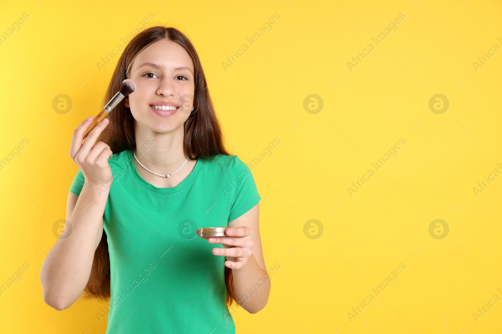 Photo of Smiling teenage girl applying blusher with makeup brush on yellow background. Space for text