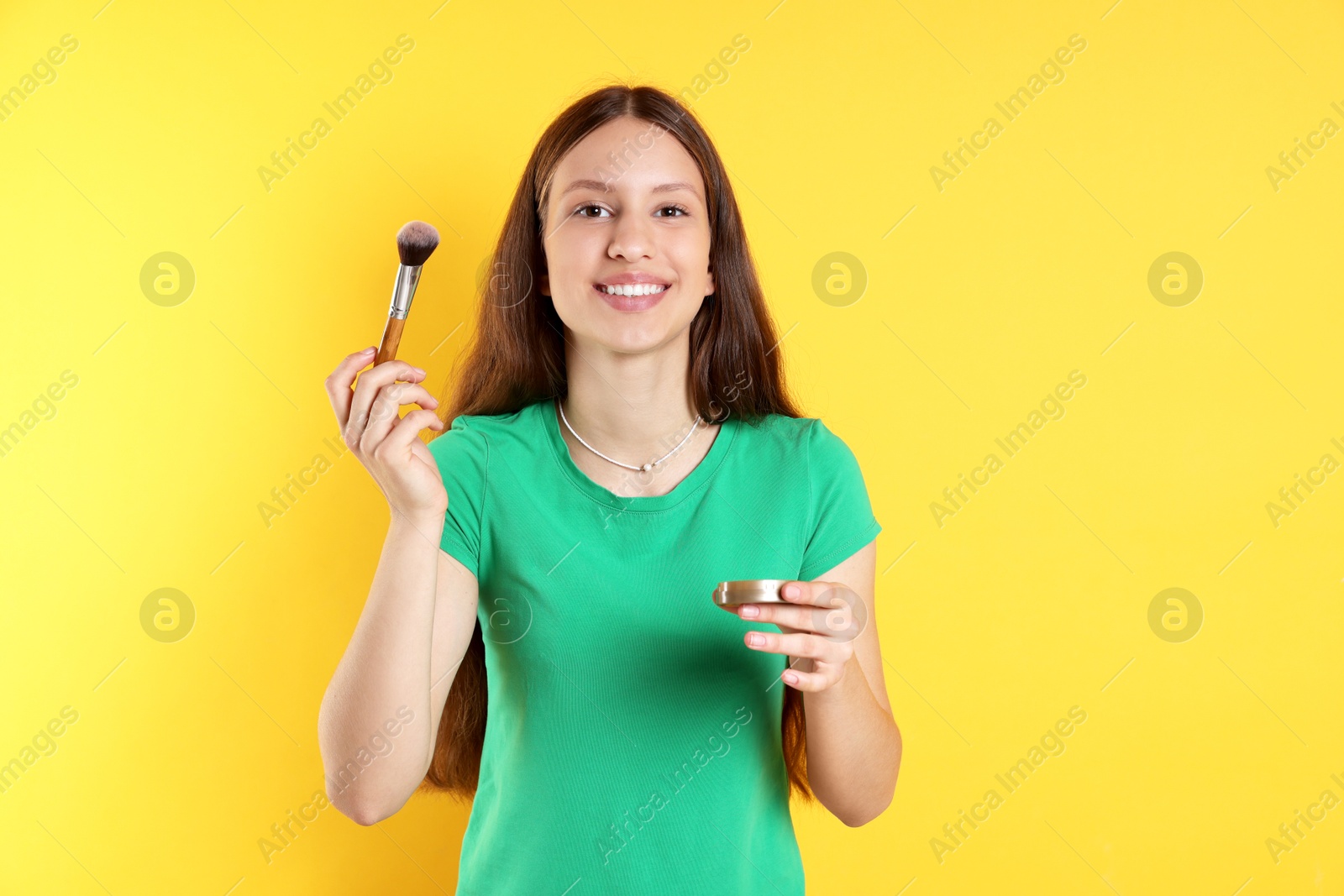 Photo of Smiling teenage girl applying blusher with makeup brush on yellow background