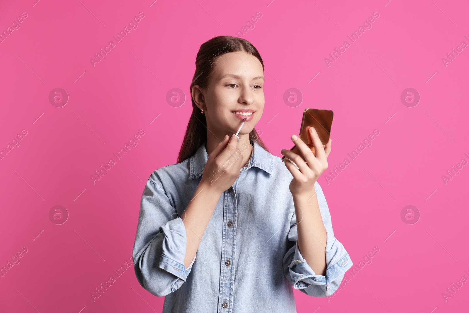 Photo of Smiling teenage girl applying lip gloss on pink background