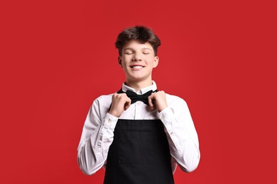 Photo of Teenage boy in apron and bowtie working as waiter on red background