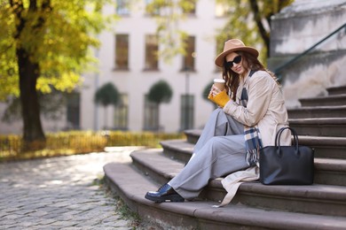 Photo of Charming young woman with cup of coffee on city street, space for text. Autumn season