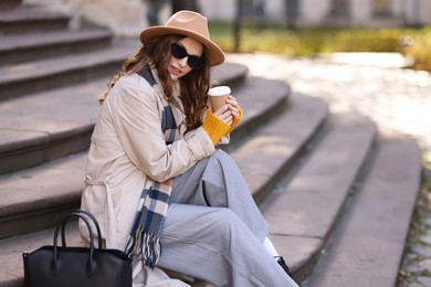 Photo of Charming young woman with cup of coffee on city street, space for text. Autumn season