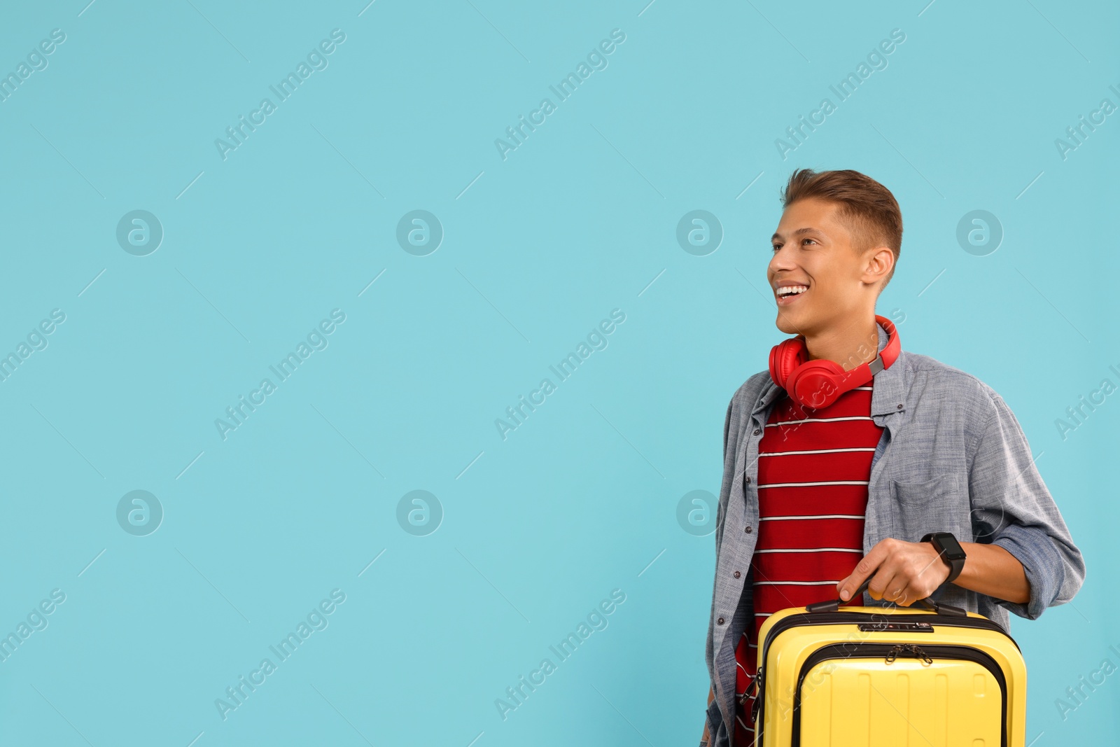 Photo of Happy traveller with headphones and suitcase on light blue background, space for text