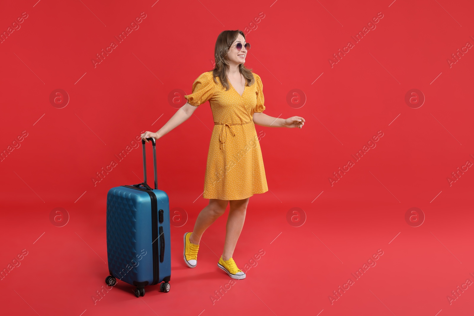 Photo of Happy traveller in sunglasses with suitcase on red background