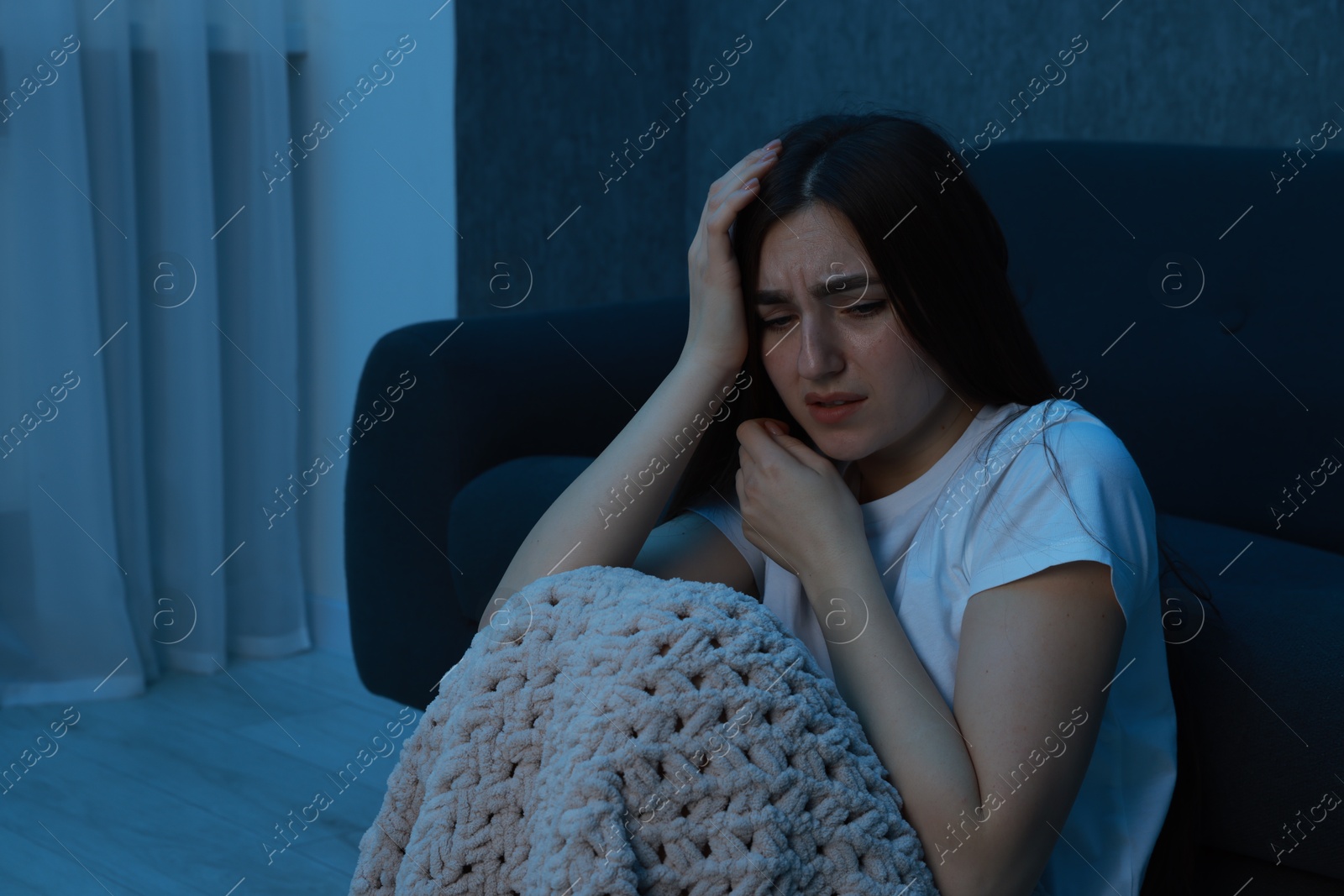 Photo of Fear of darkness. Scared young woman with blanket near couch indoors at night, space for text