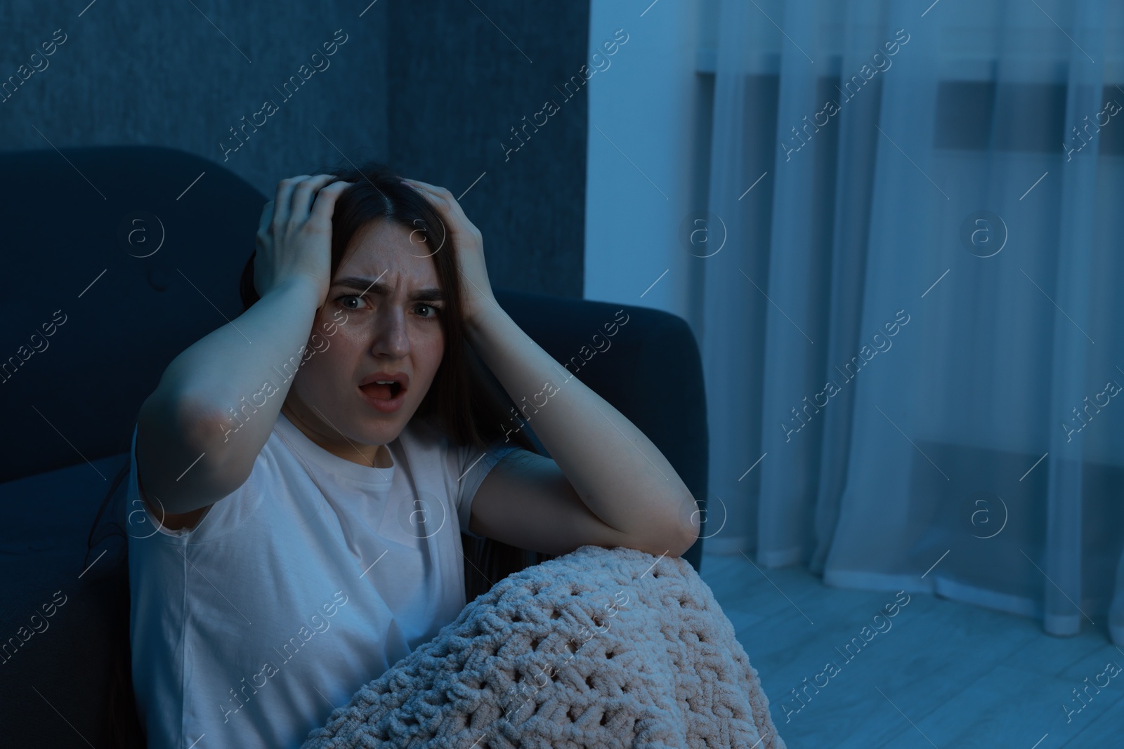 Photo of Fear of darkness. Scared young woman with blanket near couch indoors at night, space for text