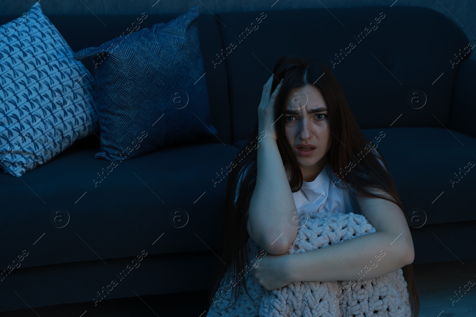 Photo of Fear of darkness. Scared young woman with blanket near couch indoors at night, space for text