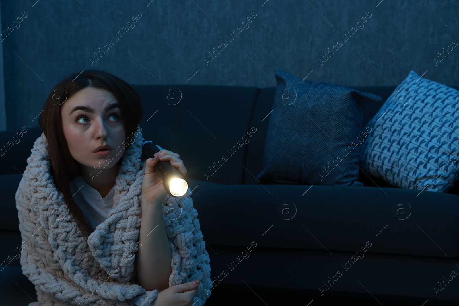 Photo of Fear of darkness. Scared young woman with flashlight and blanket near couch indoors at night, space for text