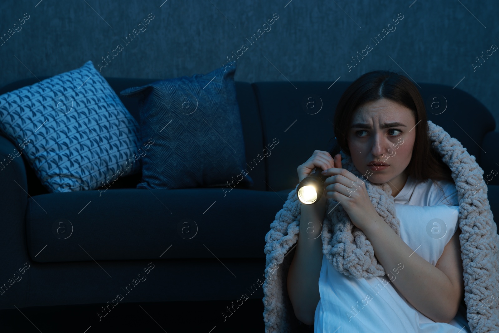 Photo of Fear of darkness. Scared young woman with flashlight hugging pillow near couch indoors at night, space for text