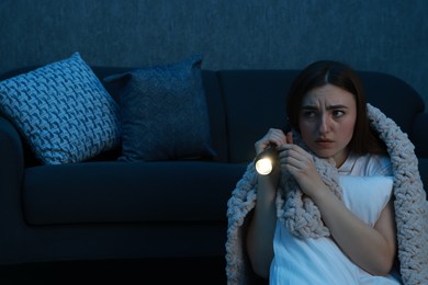 Photo of Fear of darkness. Scared young woman with flashlight hugging pillow near couch indoors at night, space for text